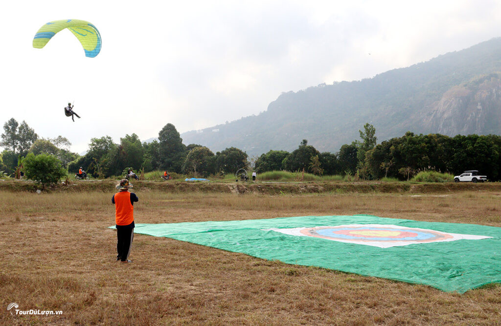 A pilot performs a precision landing at the Ho Chi Minh City Open Paragliding Tournament 2025. On the ground is a tarpaulin with a central circle as the landing spot, with a judge watching to score.