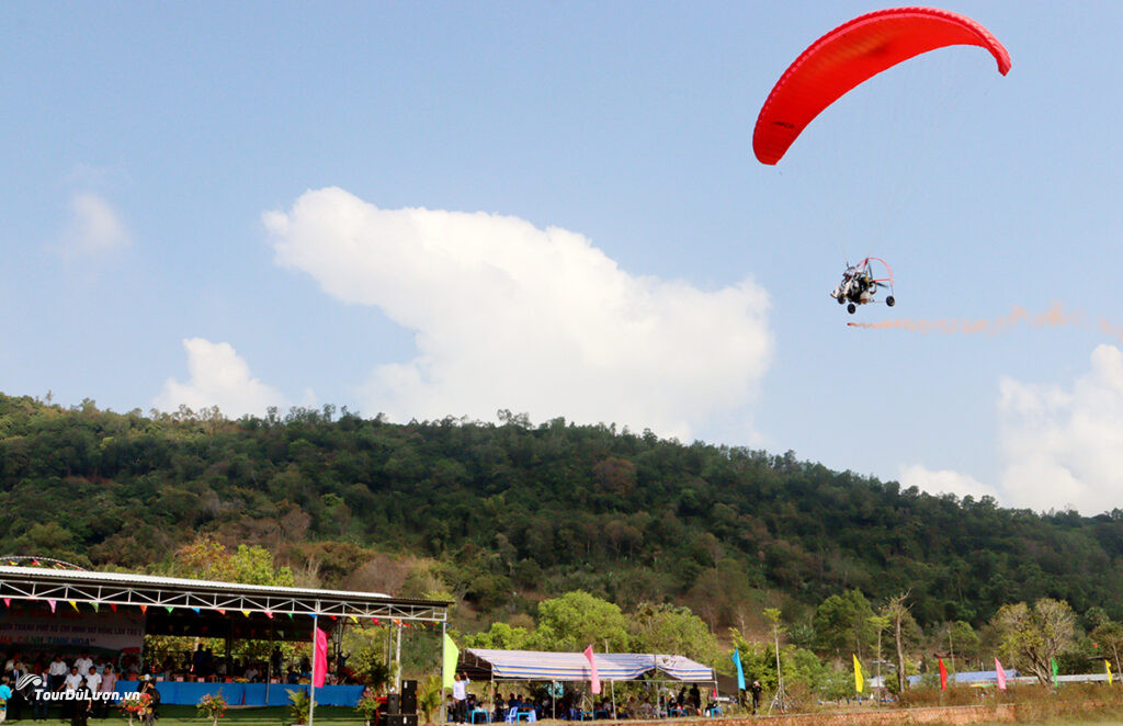 A pilot controls a powered paraglider as it flies through the air, leaving a trail of orange smoke behind. Below is the opening ceremony stage area with stands, flags and spectators.